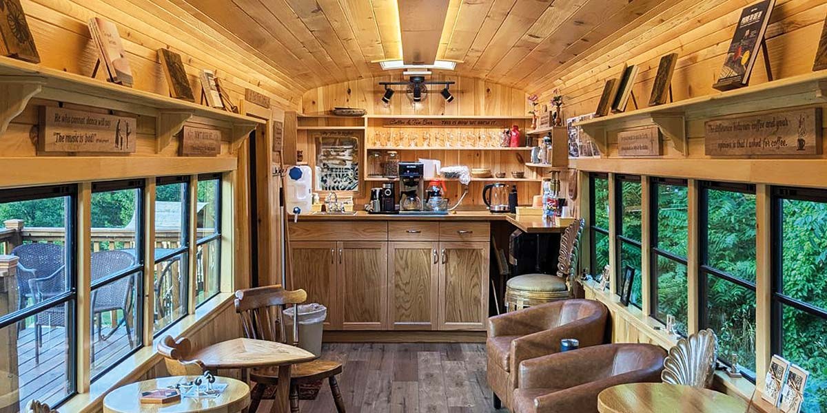 Cozy wooden cafe interior with tables, chairs, large windows, and a coffee station at the back.