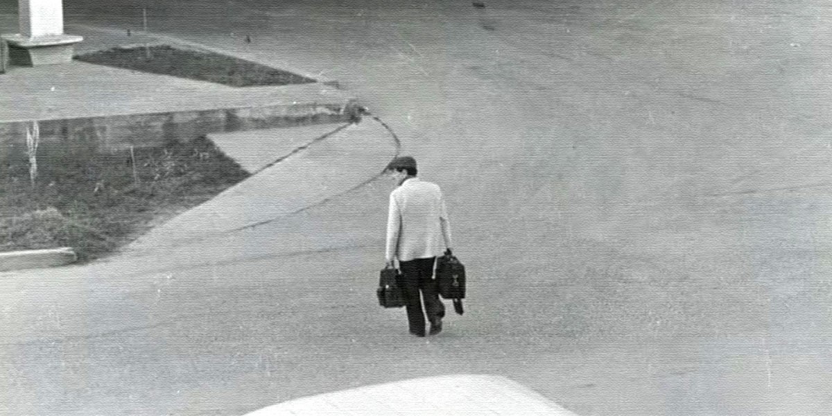 Black and white photo of a man walking on an empty street carrying two briefcases.