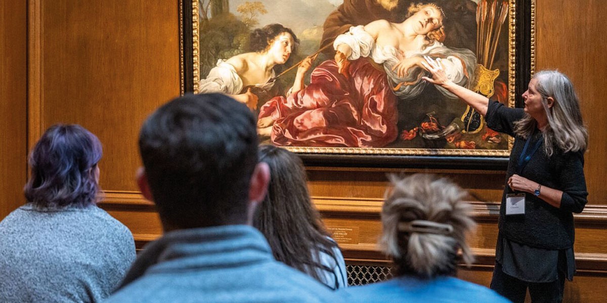 Museum guide explains a classical painting to seated visitors in a wood-paneled gallery.