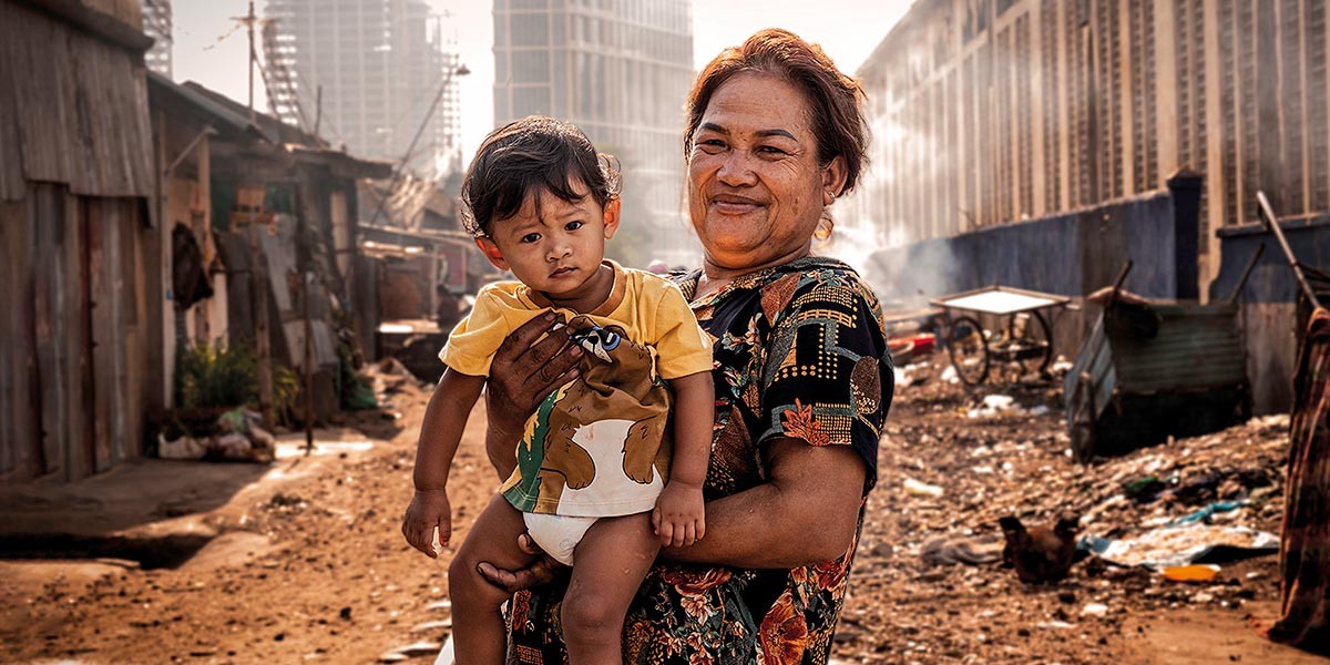 Woman holding a young child in a rundown urban area with dirt ground and buildings in the background.