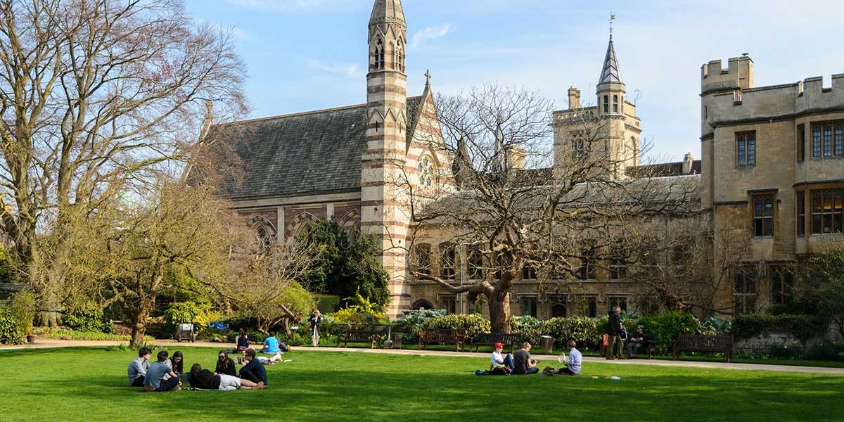 Groups of people sitting and socializing on a green lawn in front of historic stone buildings and leafless trees.