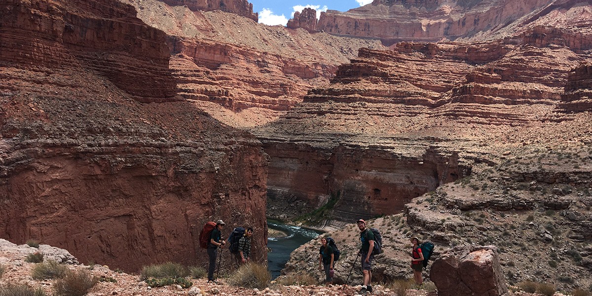 Five hikers with backpacks standing on rocky terrain near a river in a canyon under blue sky.