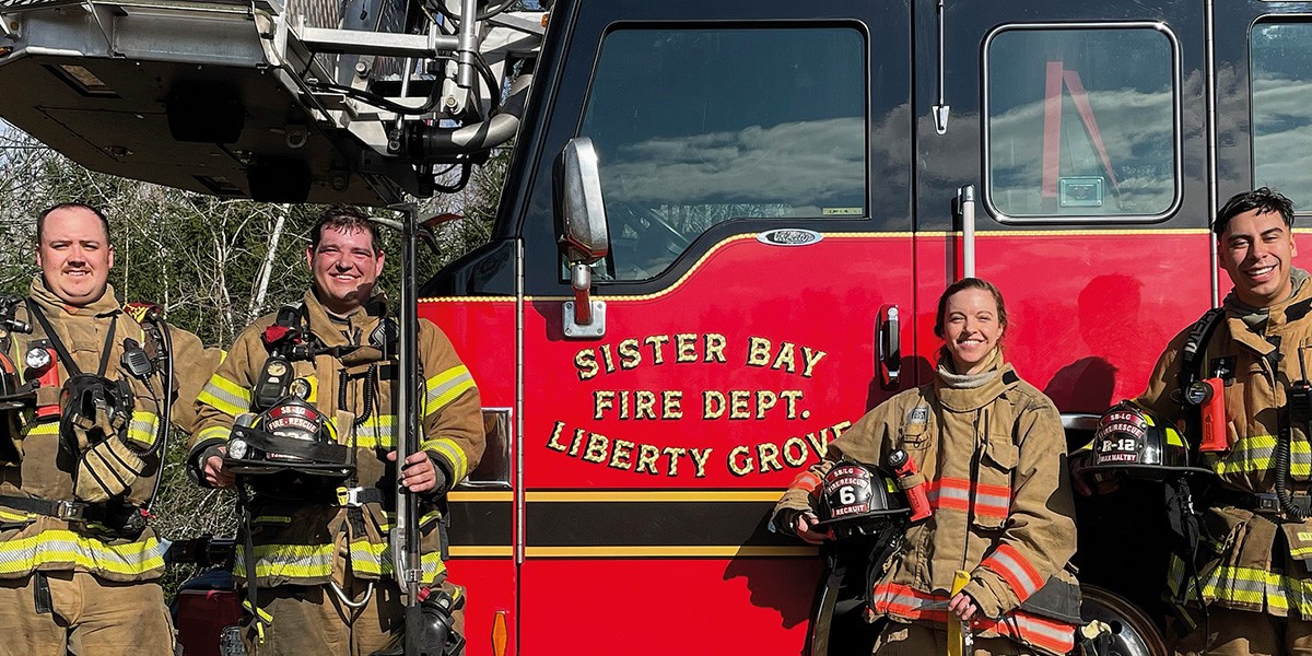 Four firefighters in gear posing in front of a red Sister Bay Fire Dept. Liberty Grove fire truck.