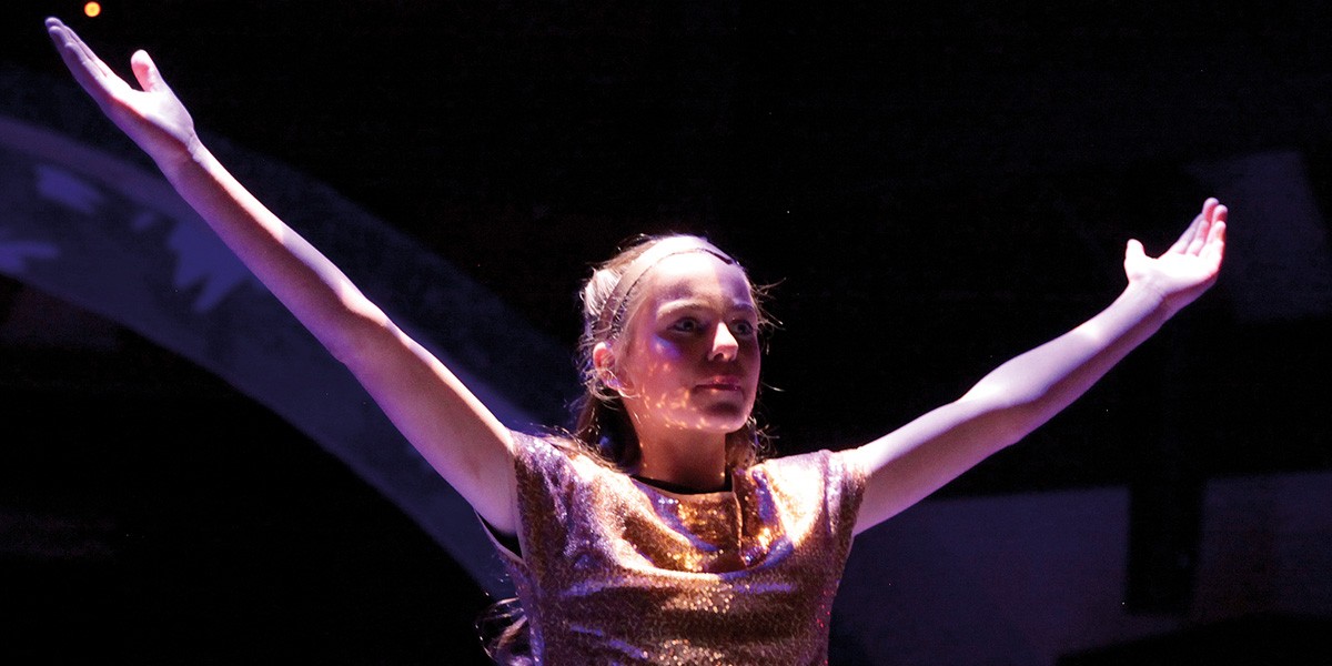 Woman in a sparkly gold top with arms raised wide on a dark stage.