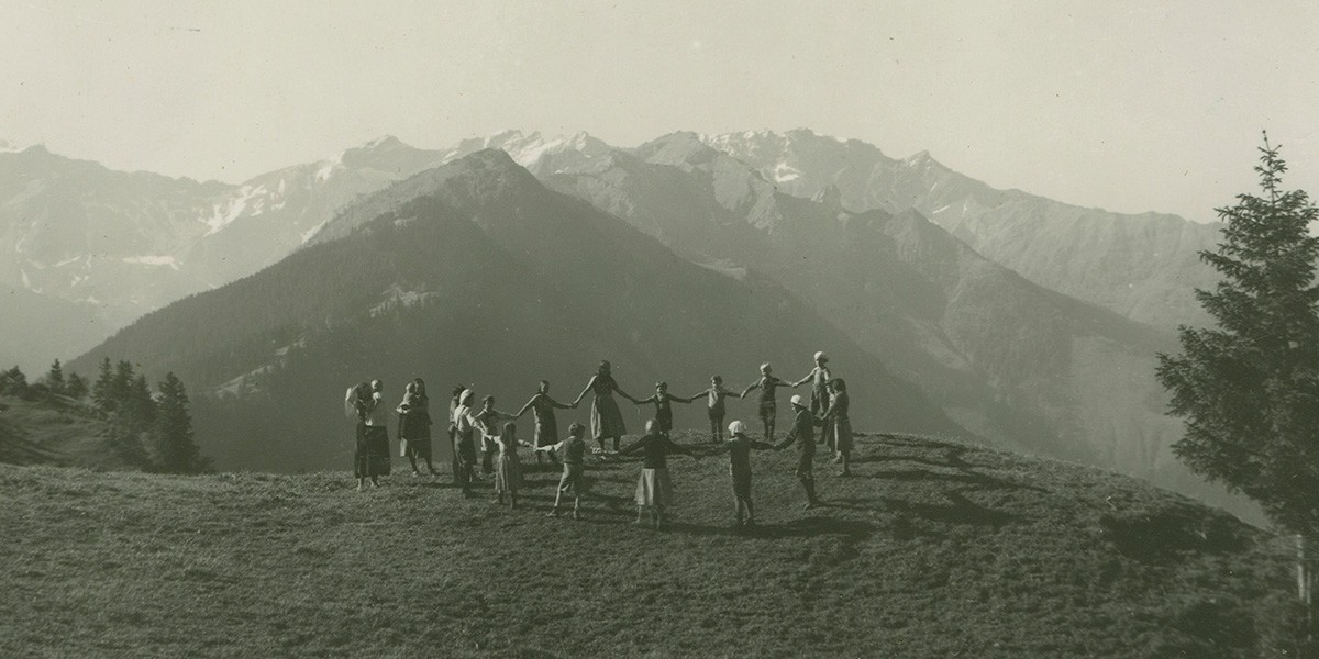Group of people holding hands in a circle on a grassy hill with mountains in the background.