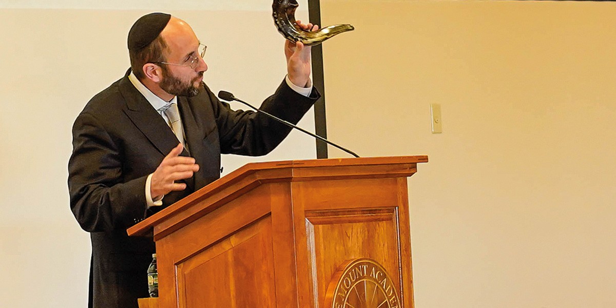 Man in suit and kippah speaking at podium, holding a shofar horn in his right hand.