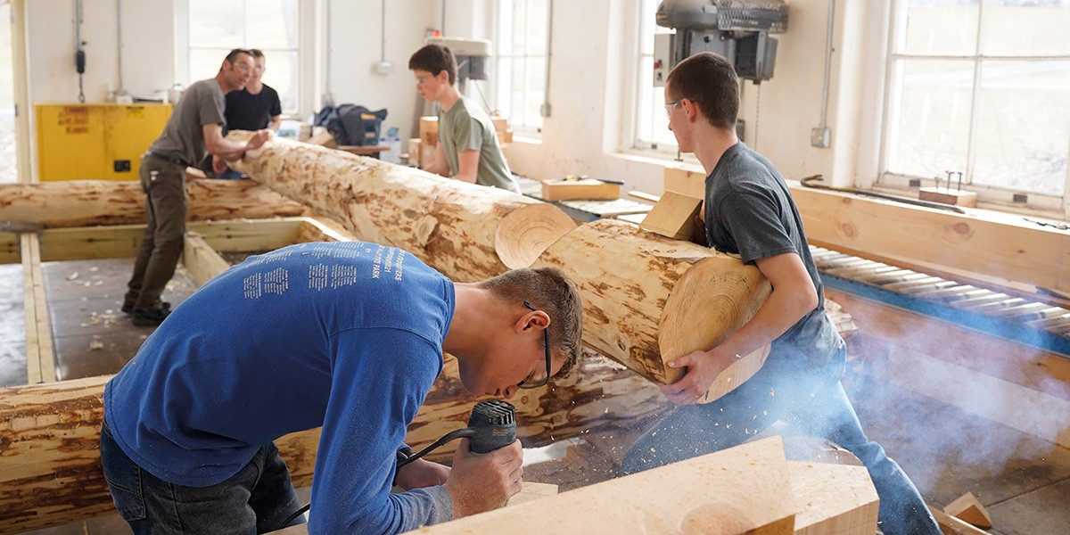 Four young men working with large logs inside a bright woodworking shop with windows.