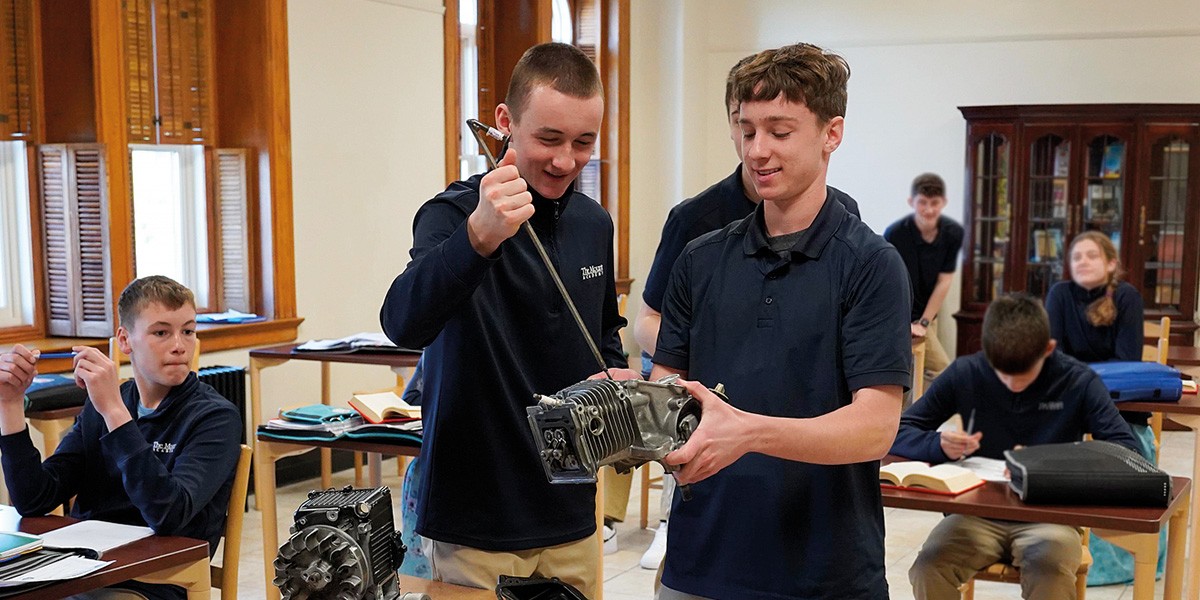 Two students in navy shirts working together on an engine part in a classroom with peers.