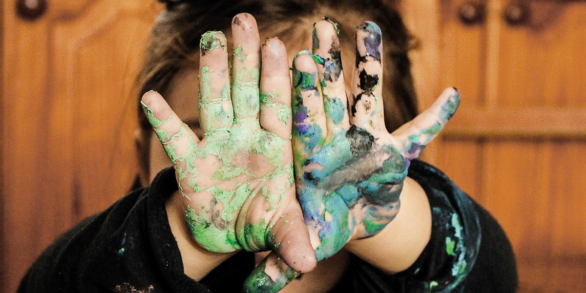 Child's hands covered in green, blue, and black paint held up in front of face.
