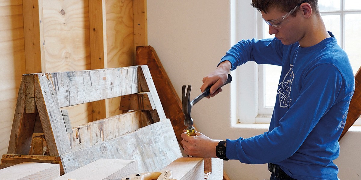 Young man wearing safety glasses hammering a nail into wood inside a workshop.