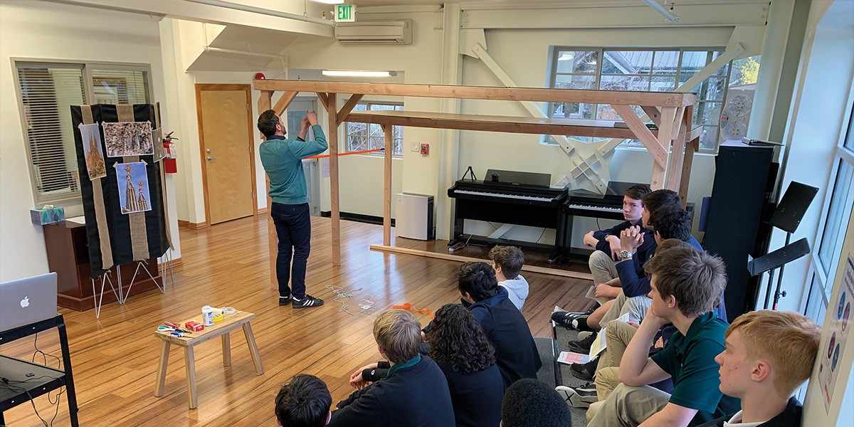 Man demonstrates weaving technique on a large wooden frame to a group of seated students in a room.