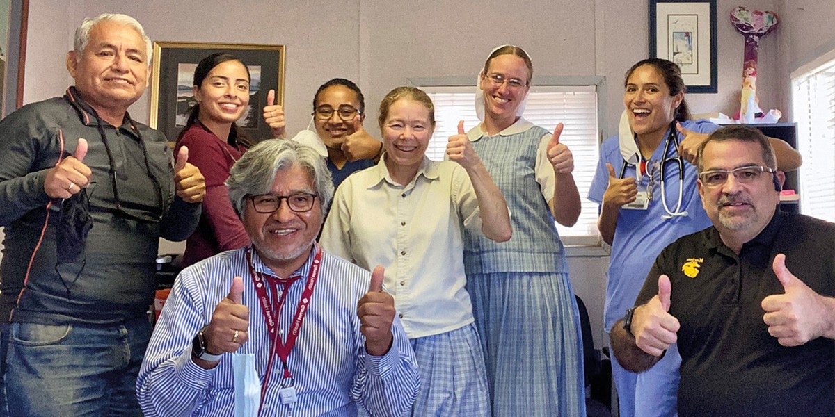 Eight diverse people indoors smiling and giving thumbs up, some wearing medical attire and glasses.