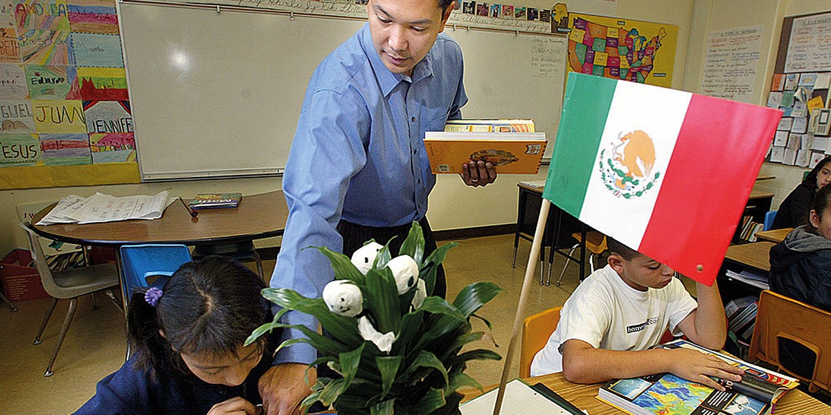 Teacher assisting young students at desks in a classroom with a Mexican flag on one desk.