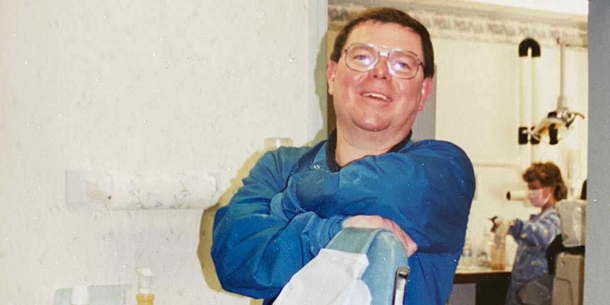 Man in blue dental scrubs leaning on chair smiling, woman in background wearing mask and dental attire.