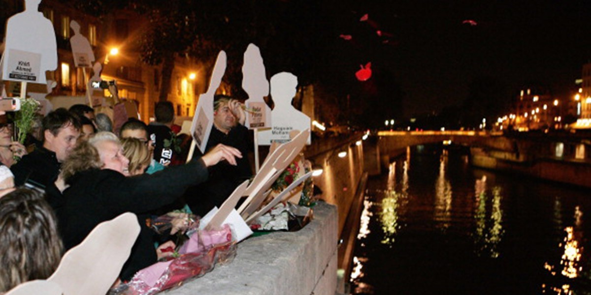 People with silhouette cutouts throwing flowers into a river at night during a vigil or memorial.