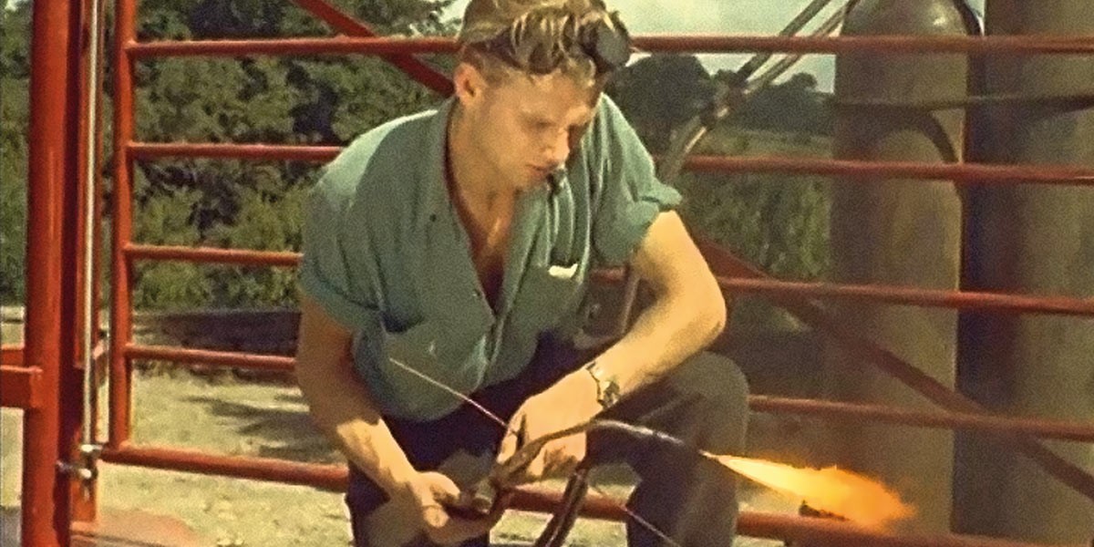 Person crouched using a blowtorch on metal near red gate outdoors in daylight.