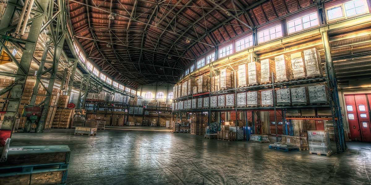 Large warehouse interior with tall shelves stacked with boxes and pallets under a wooden ceiling.