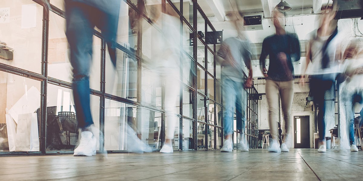 Blurred motion of people walking inside a modern office with glass partitions and wooden floors.