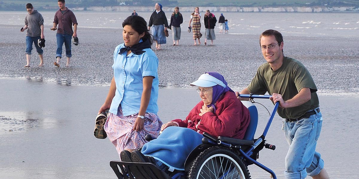 Man pushing elderly woman in wheelchair along wet beach with woman walking barefoot beside them.