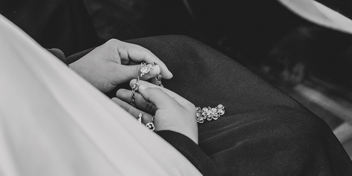 Person holding a rosary with hands resting on their lap in black-and-white photo.
