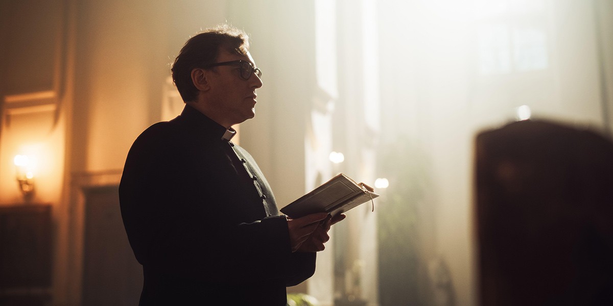 Man in clerical clothing holding an open book inside a softly lit church interior.