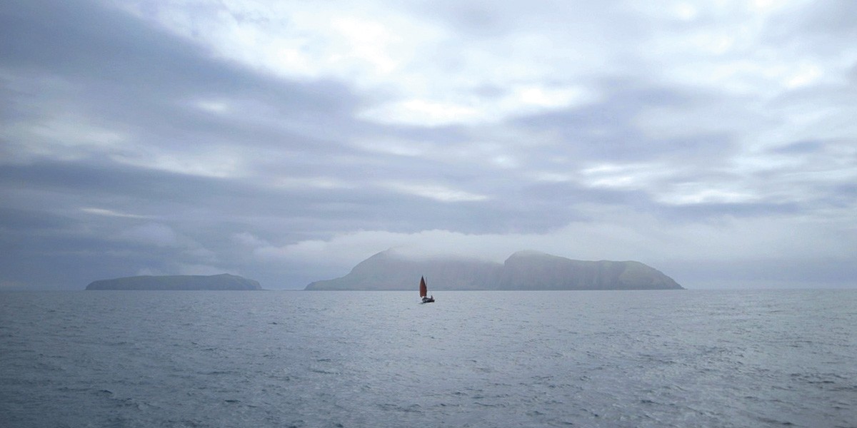 Small sailboat with a red sail on calm sea waters near distant foggy islands under cloudy sky.