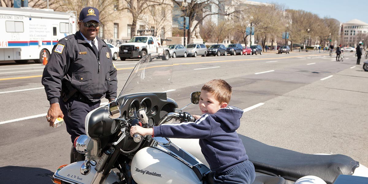 Young boy sitting on a white Harley Davidson motorcycle smiling as a uniformed police officer stands nearby.