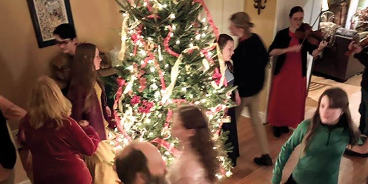 People dancing and socializing around a decorated, lit Christmas tree indoors.