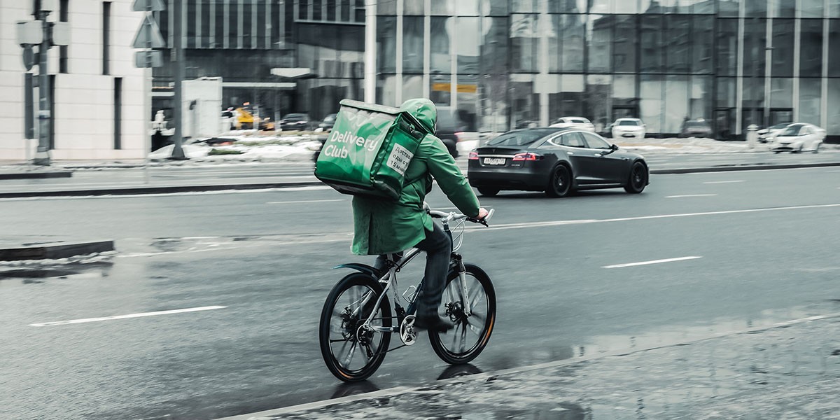 Delivery worker in green jacket riding a bicycle with green Delivery Club bag in city street traffic.