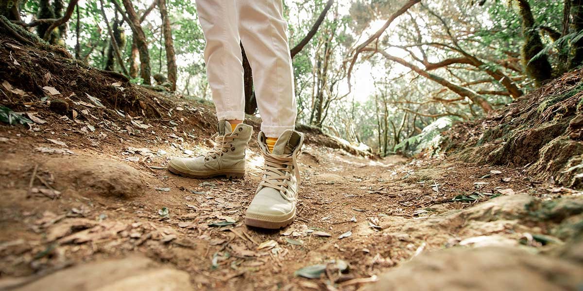 Person wearing beige boots and white pants walking on a forest dirt trail covered with leaves.