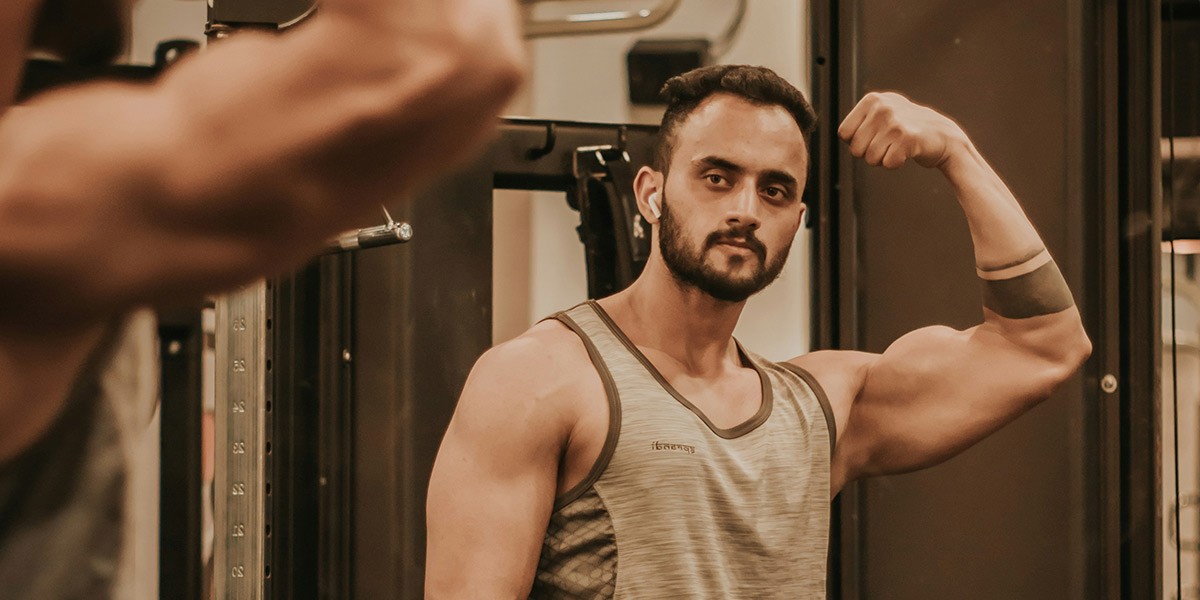 Muscular man flexing his arm and looking at himself in a gym mirror wearing a gray tank top.