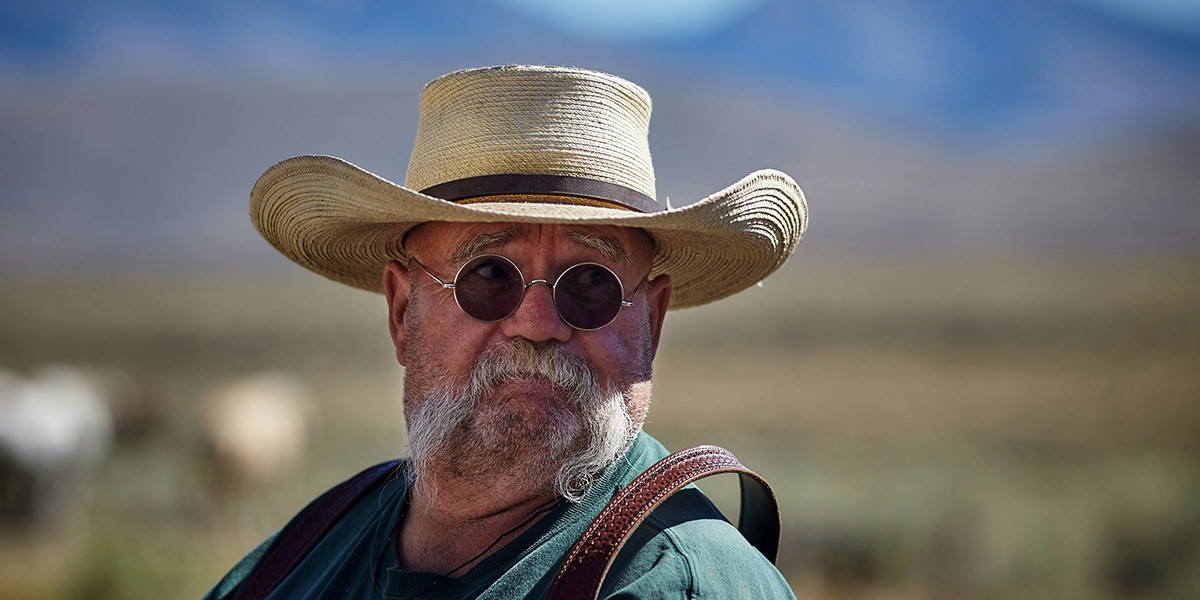 Bearded man wearing round glasses and straw hat outdoors with blurred landscape background.