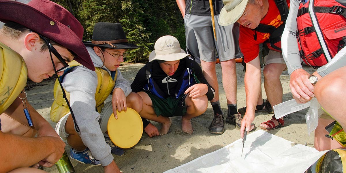Group of hikers gathered on sand studying a map while one points with a knife, wearing hats and backpacks.