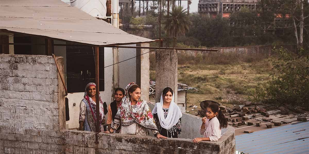 Five women and girls in traditional clothing standing on a rooftop balcony in a rural area.