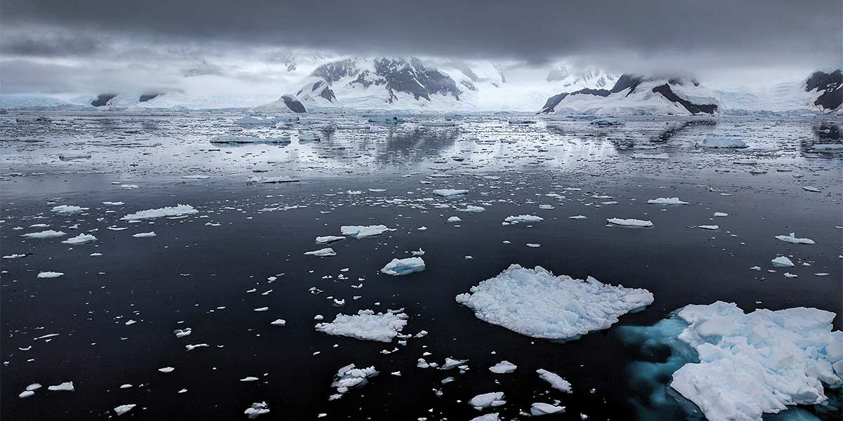 Cold ocean scattered with ice chunks beneath dark, heavy clouds with snowy mountains in the background.