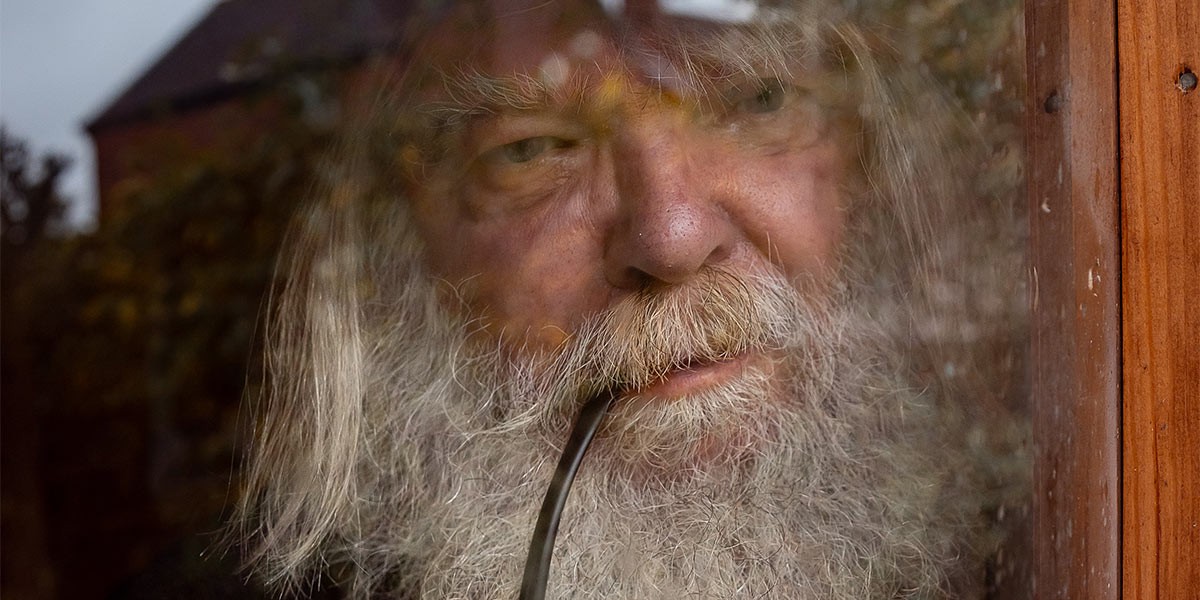 Older man with a long white beard smoking a pipe, seen through a window with reflections.