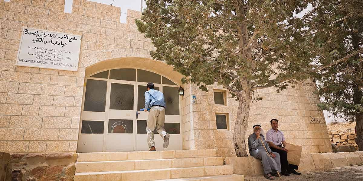 A man approaches a beige stone building entrance while two men sit on a nearby stone bench under trees.