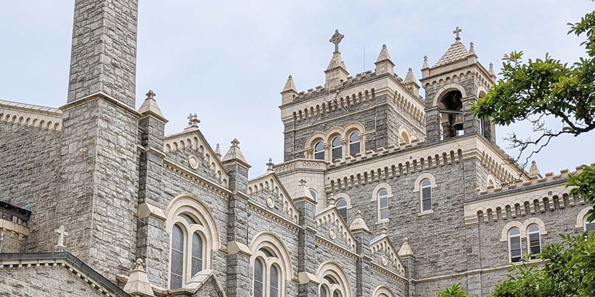 Stone church tower with ornate architecture, arched windows, crosses, and greenery in the foreground.