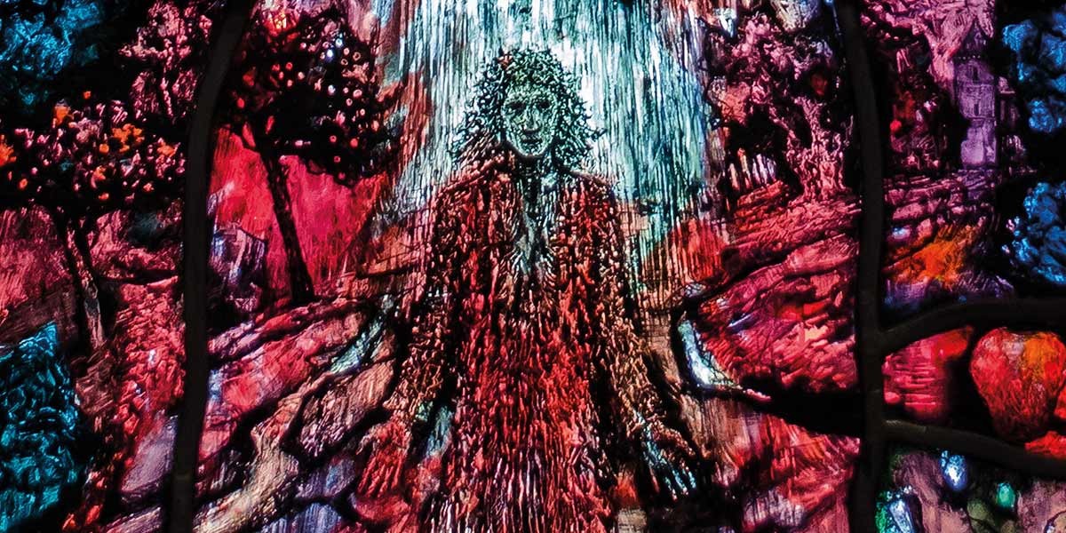 Stained glass artwork depicting a figure with flowing hair in a red robe surrounded by rocks and trees.