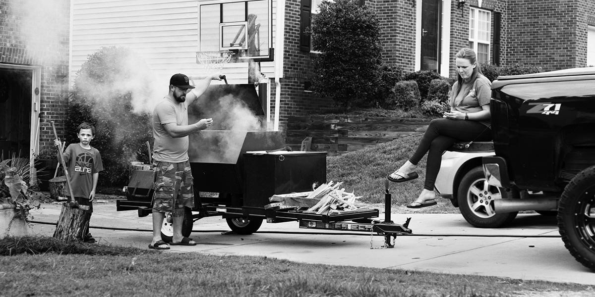 Man tending a smoking barbecue grill while boy and woman nearby in a driveway outside a brick house.