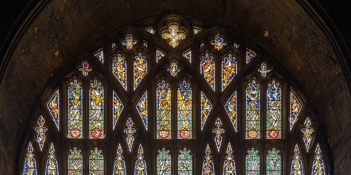 Arched stained glass window with colorful religious figures and angel motifs in a stone building.