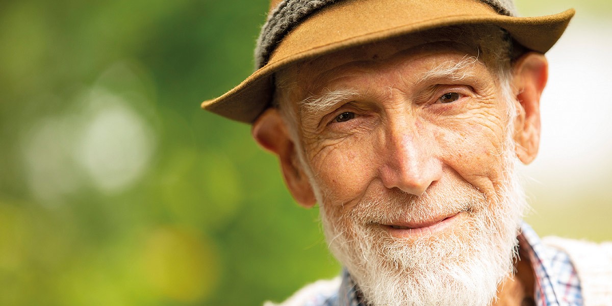 Close-up of an elderly man with a white beard wearing a brown hat and smiling gently outdoors.