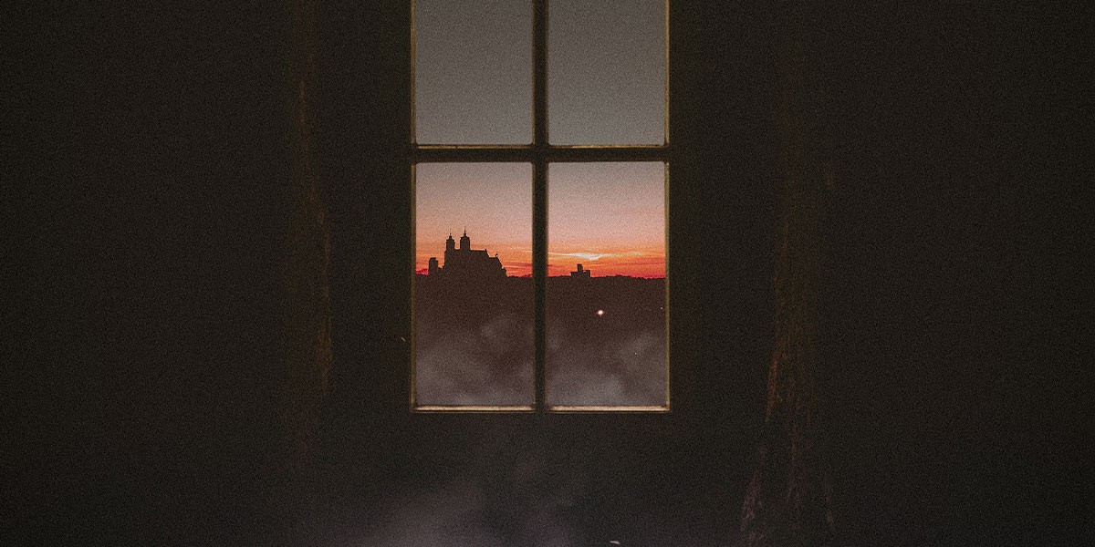 Sunset sky with orange and pink hues seen through four-paned window with dark interior walls.