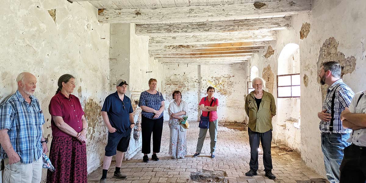 Group of nine people standing and listening inside a rustic room with exposed white beams and peeling walls.