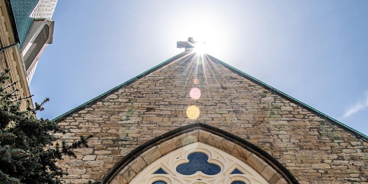 Sunlight shining behind a cross atop a stone church roof with a pointed window beneath.