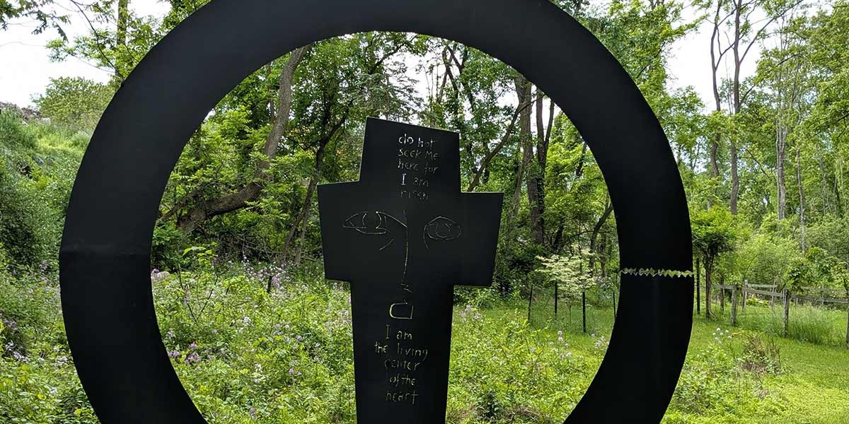 Black circular sculpture outdoors framing a cross shape with engraved eyes and text, surrounded by greenery.