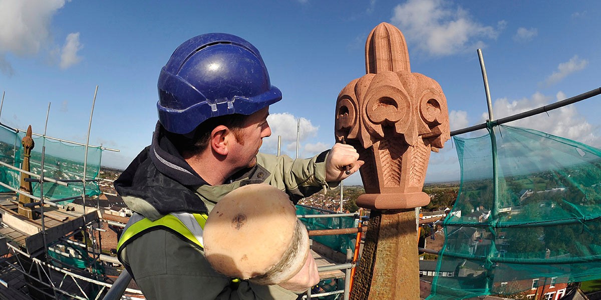 Man wearing a blue helmet and safety vest carving ornate stonework on building scaffolding outdoors.
