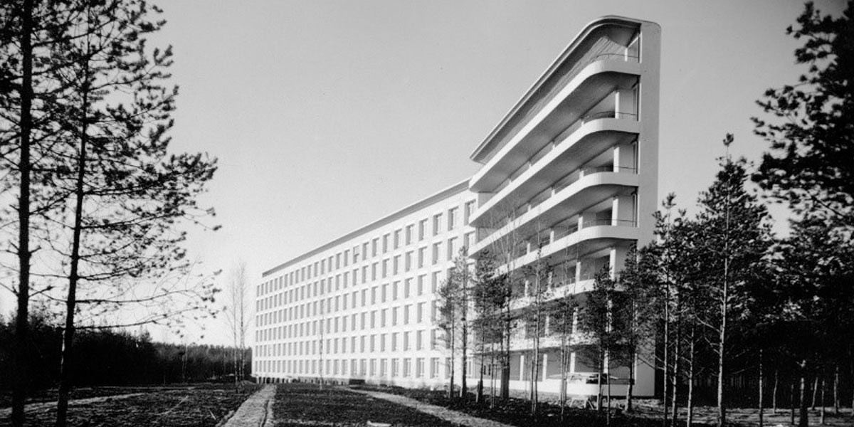 Long modern building with curved balconies surrounded by tall thin trees in a sparse forest.