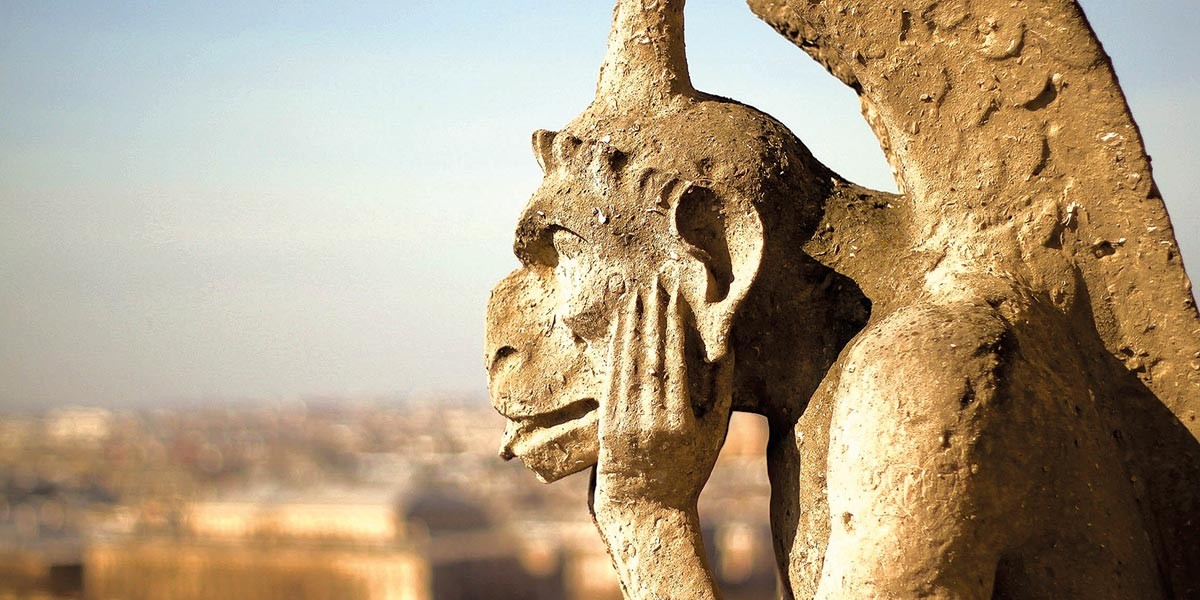Side view of a stone gargoyle resting its head on its hand with a blurred cityscape background.