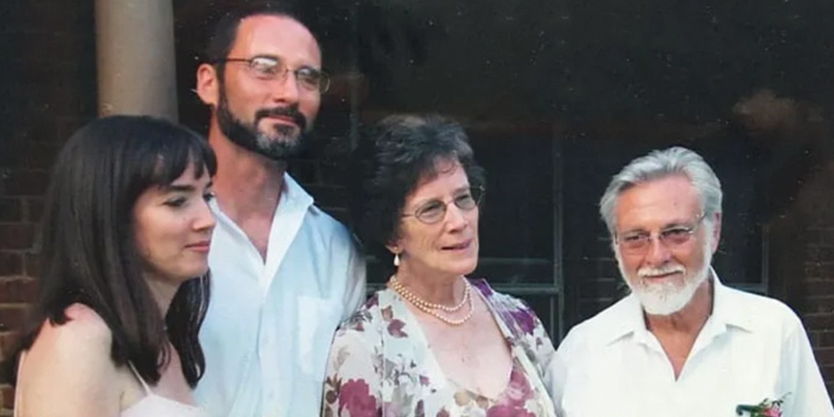 Four adults standing indoors, three wearing glasses, dressed in light-colored clothing.